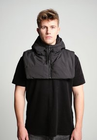 Vesta puffer neagră cu guler înalt și fermoar frontal, având un design căptușit. Pur tată pe un tricou negru, estetică minimalistă.