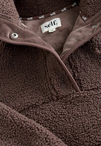 Pullover in pile marrone con una texture morbida, caratterizzato da un sistema di chiusura a scatto sul colletto e un'etichetta cucita all'interno del collo.