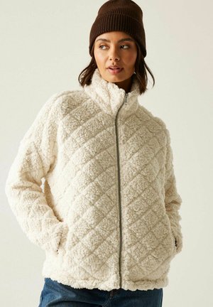 Femme portant une veste en polaire matelassée crème, les mains dans les poches, avec un bonnet en maille marron, regardant sur le côté sur un fond uni.