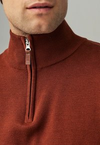 Svetr s čtvrtinovým zipem v rezavé barvě, s žebrovanou texturou a vysokým límcem. Obsahuje stříbrný zip a malý logo detail na táhle.