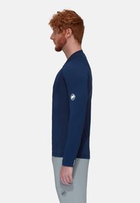 Dunkelblaues Langarmshirt mit strukturiertem Design und weicher Textur. Mit einem Logo auf dem linken Arm und taillierten Ärmeln. Kombiniert mit hellgrauen Hosen.
