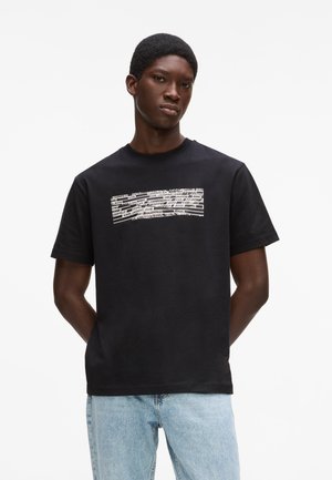 Junger Mann trägt ein schwarzes T-Shirt mit einem weißen rechteckigen Motiv, auf dem mehrmals der Schriftzug "Karl Lagerfeld Jeans" abgebildet ist, kombiniert mit hellblauen Jeans.