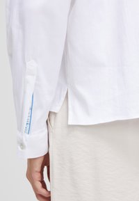 White long-sleeve shirt with a button cuff, featuring a colorful inner lining at the cuff and side slits; smooth, lightweight fabric.
