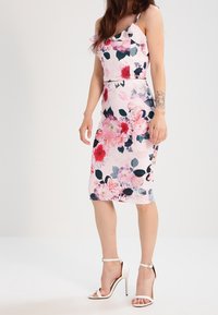 Vestido midi floral en tonos rosas con diseño asimétrico de volantes en un solo hombro, que presenta estampados vivos de peonías y hojas verde oscuro. Combinado con tacones blancos.
