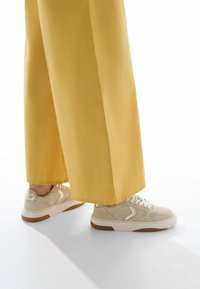 Beige sneakers with knit panels, white laces, and textured soles, paired with wide-leg yellow trousers. Light background.