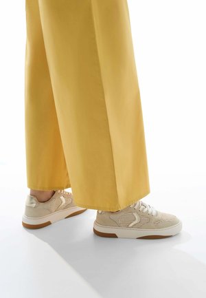 Sneaker beige con pannelli in maglia, lacci bianchi e suole testurizzate, abbinati a pantaloni gialli a gamba larga. Sfondo chiaro.