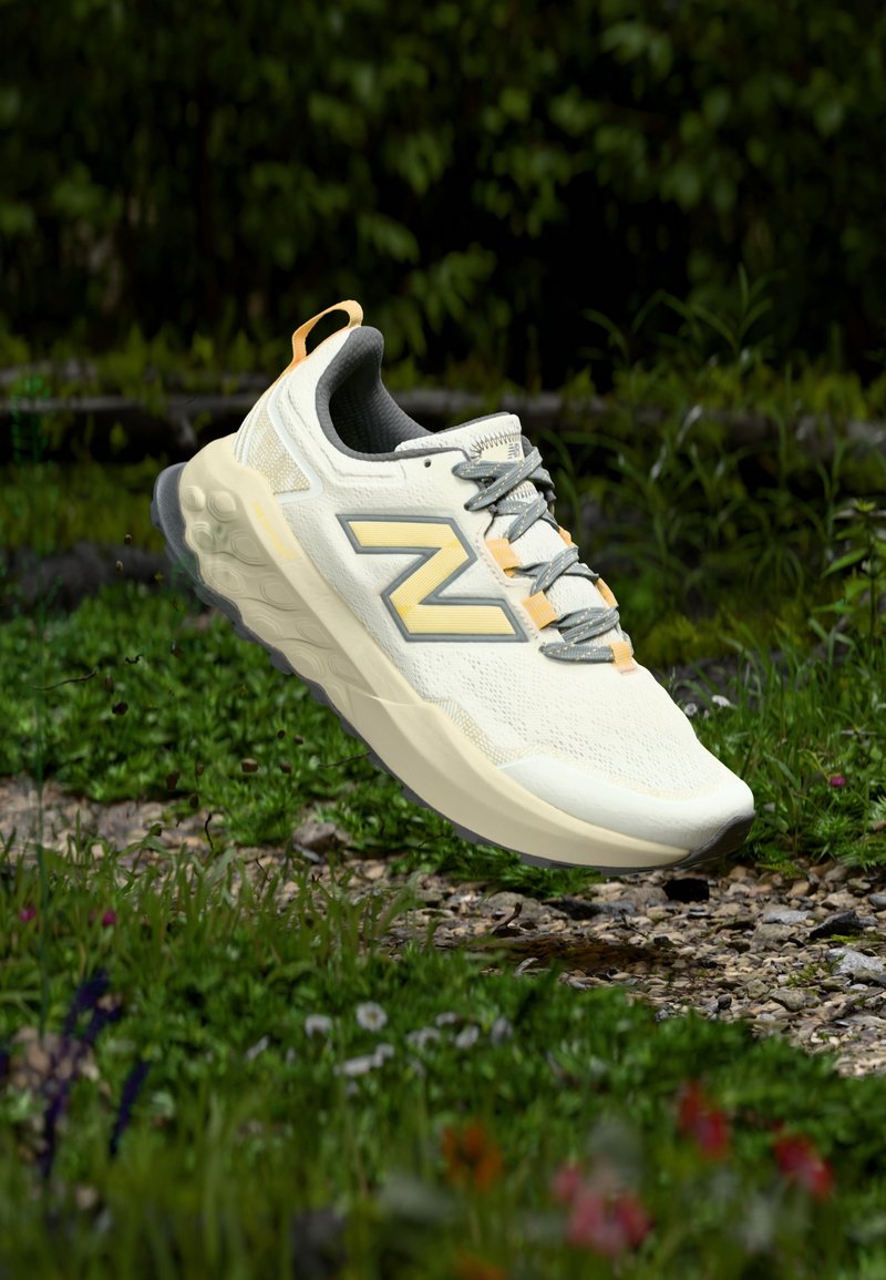 Chaussure de sport beige clair et grise avec une semelle épaisse, flottant au-dessus d'un chemin de gravier entouré d'herbe et de petites fleurs.