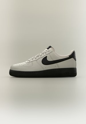 Baskets basses blanches avec le swoosh Nike noir, languette de talon noire et semelle noire épaisse sur un fond neutre.