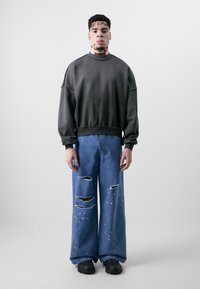 Sweatshirt surdimensionné gris foncé avec des poignets côtelés, associé à un jean large bleu clair comportant des déchirures et des accents blancs éparpillés.