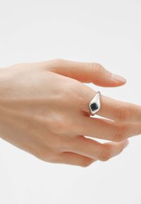 Anillo de plata con un diseño suave y cónico, que presenta una gran piedra cuadrada oscura engastada en la parte superior, llevado en una mano de piel clara.