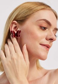 Crvene naušnice u obliku leptira s slojevitim laticama, s sjajnim finišem i kontrastnim svjetlijim rubovima, nošene na uhu osobe.