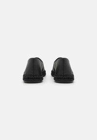 Des chaussures noires à enfiler avec une tige en cuir lisse et des détails en corde tressée autour de la semelle. Forme arrondie, design minimaliste, sans matériel visible.