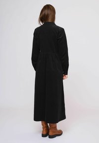 Rochie din catifea neagră cu mâneci lungi, un corp ajustat și o talie evazată, asociată cu cizme maro până la genunchi. Detaliu de guler simplu.