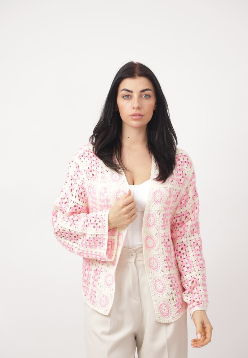 Mujer de cabello oscuro que lleva una blusa y pantalones blancos, sosteniendo abierta una chaqueta de crochet en tonos rosa claro y blanco sobre un fondo liso.