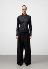 Top lungo a maniche lunghe nero e texturizzato con colletto alto, abbinato a pantaloni larghi neri. Il modello indossa un anello d'argento e scarpe nere.