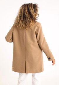 Mujer con cabello rizado vestida con un abrigo largo beige con detalles de botones negros y pantalones blancos, de espaldas contra un fondo blanco.