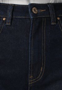 Mörkblå denimjeans har en knappstängning, kontrasterande gul sömnad och sidofickor. Tyget har en slät, lätt texturerad yta.