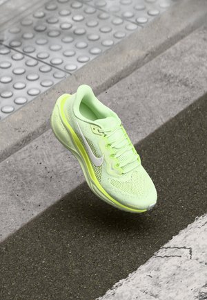 Chaussure de course Nike vert clair et jaune flottant au-dessus d'un trottoir texturé et d'une route asphaltée avec une ligne blanche peinte.