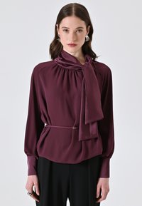 Blusa borgogna con scollo drappeggiato, dettaglio del fiocco, maniche lunghe e vita scambiata con cintura; tessuto morbido e liscio con una leggera lucentezza.