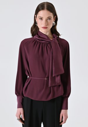 Blusa borgogna con scollo drappeggiato, dettaglio del fiocco, maniche lunghe e vita scambiata con cintura; tessuto morbido e liscio con una leggera lucentezza.
