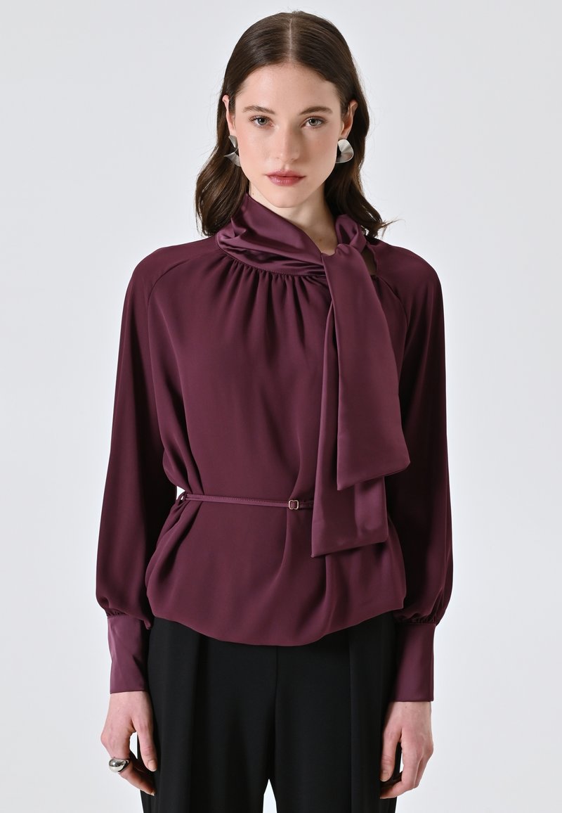Blusa borgogna con scollo drappeggiato, dettaglio del fiocco, maniche lunghe e vita scambiata con cintura; tessuto morbido e liscio con una leggera lucentezza.