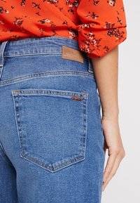 Jeans de mezclilla azul con cintura alta, que cuentan con una etiqueta de cuero en la parte posterior y un solo bolsillo visible en el lado derecho.