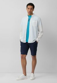 Lichtblauw overhemd over een teal t-shirt, marineblauwe shorts en witte sneakers. Zachte texturen en eenvoudige, strakke lijnen overal.