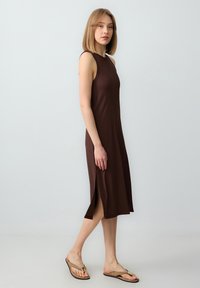 Brązowa sukienka bez rękawów z bocznymi rozcięciami, wykonana z prążkowanej tkaniny, o dopasowanym kroju. Modelka połączyła ją z beżowymi klapkami.
