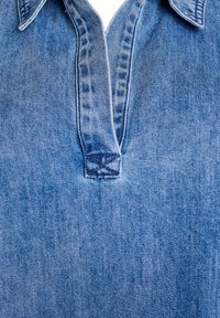 Denimskjorte med kraveudskæring, lyseblå farve, diskret falmet tekstur og en lille syet detalje ved kraven.