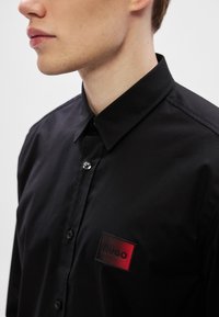 Μαύρο πουκάμισο με κουμπιά και μυτερό γιακά, με υφασμάτινο λογότυπο HUGO σε κόκκινο χρώμα στο στήθος. Λείο ύφασμ�α, εφαρμοστή σχεδίαση.