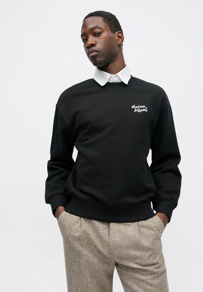 Čierna mikina s rebrovanými manžetami a zakončením, s bielou vyšitou značkou "Maison Kitsuné" na ľavej hrudi, nosená cez bielu košeľu s golierom.