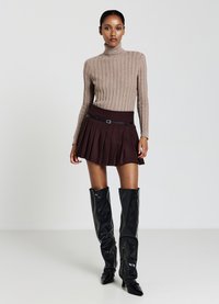 Beige gerippter Rollkragenpullover, braider Faltenminirock mit einem Gürtel und glänzende schwarze kniehohe Stiefel mit spitzen Zehen.