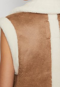 Brun imitationsmockaväst med krämfärgad imitationspälsfoder, med en ren kant i halslinjen och sömlös design för en mysig, lager-på-lager-look.