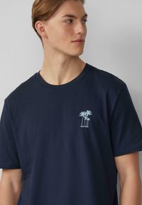 Marineblauwe katoenen t-shirt met een kleine lichtblauwe palmboomafbeelding en de tekst "STAY COOL" geborduurd op de borst.