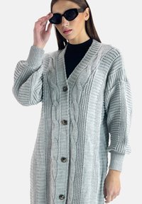 Ljust grått stickat cardigan med flätmönster, ribbad textur och v-ringning. Har stora bruna knappar och överdimensionerade ärmar.