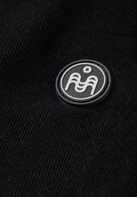 Musta samettikangasta, jossa on pyöreä mustavalkoinen logomerkki, jossa on aaltoilevia viivoja ja piste niiden yläpuolella. Pintakuvio on uritettu ja kosketeltava.