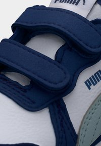 Λευκό αθλητικό παπούτσι με ναυτικά μπλε και ανοιχτά γκρι στοιχεία, διαθέτει συνθετικό επάνω μέρος, ιμάντες βέλκρο και υφή στη σόλα. Ορατό το λογότυπο PUMA.