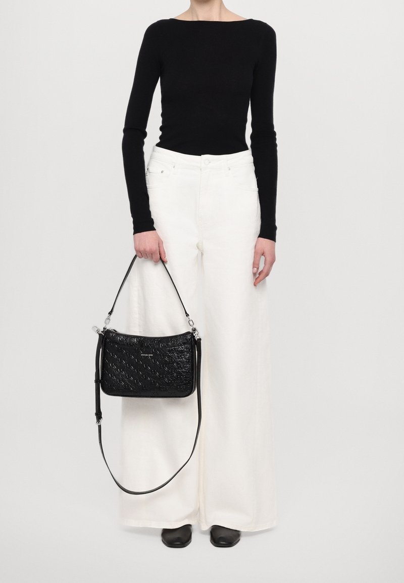 Personne portant un haut noir à manches longues et un pantalon blanc à jambes larges, tenant un sac à main noir texturé avec une bandoulière.