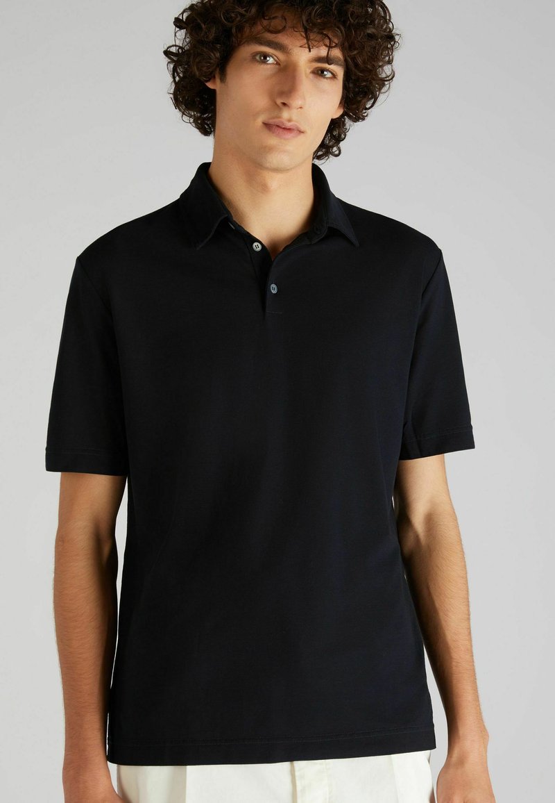 Camisa polo negra de tela suave, con mangas cortas, un cuello plano y una tapeta de tres botones, con un diseño minimalista y sin patrones.
