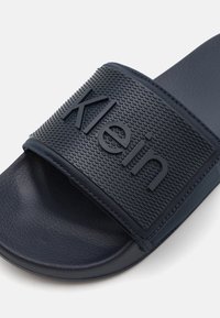 Tengerészeti kék csúszó szandál, melynek texturált gumiszíja van, rajta az "Klein" felirattal. A talpa sima felületű, kényelmet biztosít.