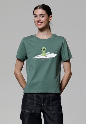 T-shirt vert à manches courtes avec un graphique d'un alien cartoon sur un avion en papier blanc, se détachant sur un fond étoilé subtil.