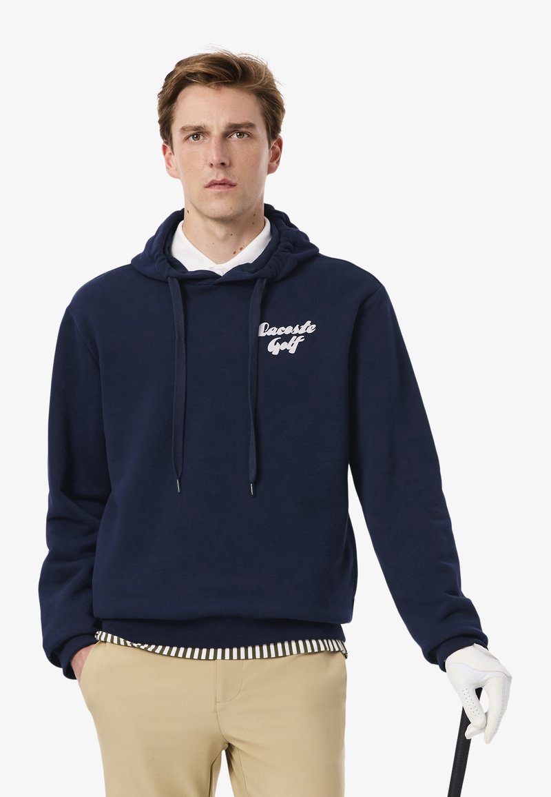 Felpa blu navy in pile con coulisse e logo "Lacoste Golf". Presenta un look a strati con una camicia colletto bianca sotto.
