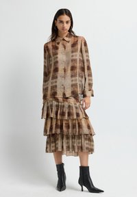 Camisa de botones de color marrón con un diseño estampado, combinada con una falda de volantes en capas. El conjunto presenta una tela suave y fluida, y está complementado con botas negras de tobillo.
