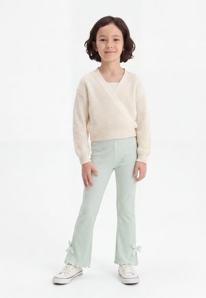 Παιδί με κοντά σκούρα μαλλιά που φοράει κρεμ πουλόβερ πλεκτό, ανοιχτό πράσινο ριμπ παντελόνι με φιόγκους στους αστραγάλους και άσπρα αθλητικά παπούτσια, όρθιο.