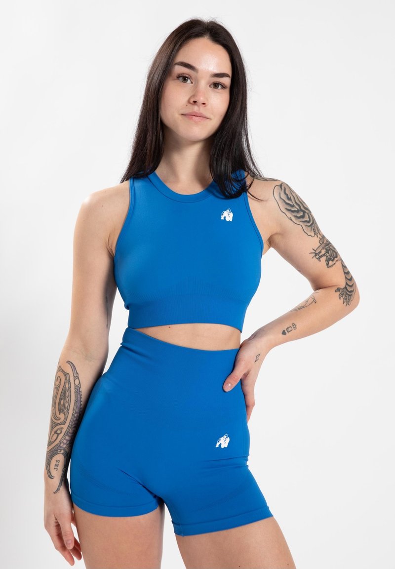 Blauwe sportieve crop top met een ronde hals en uitsnijding in de taille, gedragen met hoge taille shorts. Beide hebben een klein wit logo. Glad textuur.