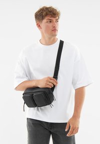 Svart axelväska med två dragkedjefack, tillverkad av mjukt syntetmaterial, med en justerbar rem och minimalistisk design.