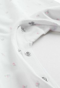 Hvid stof onesie med blomstermønster i lyserød. Trykknapper nær halsen. Blødt, elastisk materiale med en glat tekstur.