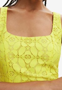 Top de cuello cuadrado de encaje amarillo brillante con patrones florales; el tejido texturizado revela la piel debajo, destacando los detalles del diseño.