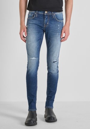 Man draagt vervaagde blauwe slim-fit jeans met verweerde details en zwarte enkellaarzen, staand tegen een effen achtergrond.