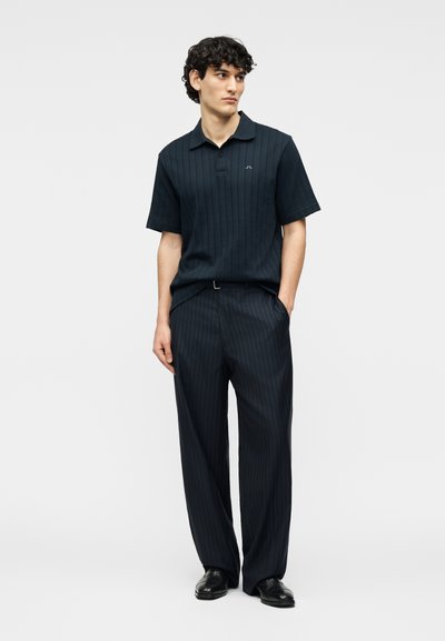 Άνδρας που φοράει μαύρο πόλο με κοντά μανίκια και γιακά και φαρδιά μαύρα υφασμάτινα παντελόνια με λεπτές ρίγες, με μαύρα επίσημα παπούτσια, στέκεται με τα χέρια στις τσέπες.